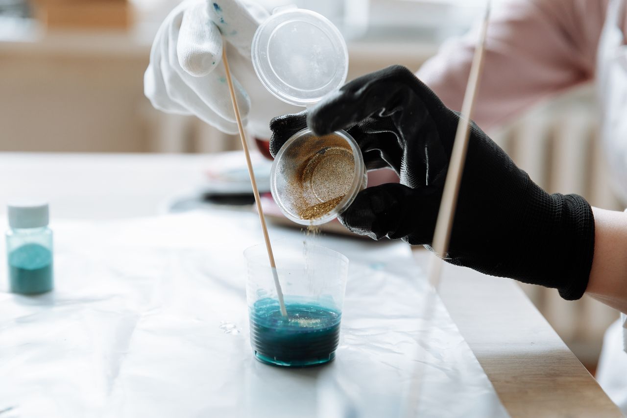 Hands in gloves mix blue and gold resin in a cup, showcasing art creation process.